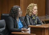 Representative Richardson sits at the testifiers' table and speaks to someone out of view. Rebecca Wallin sits next to her.